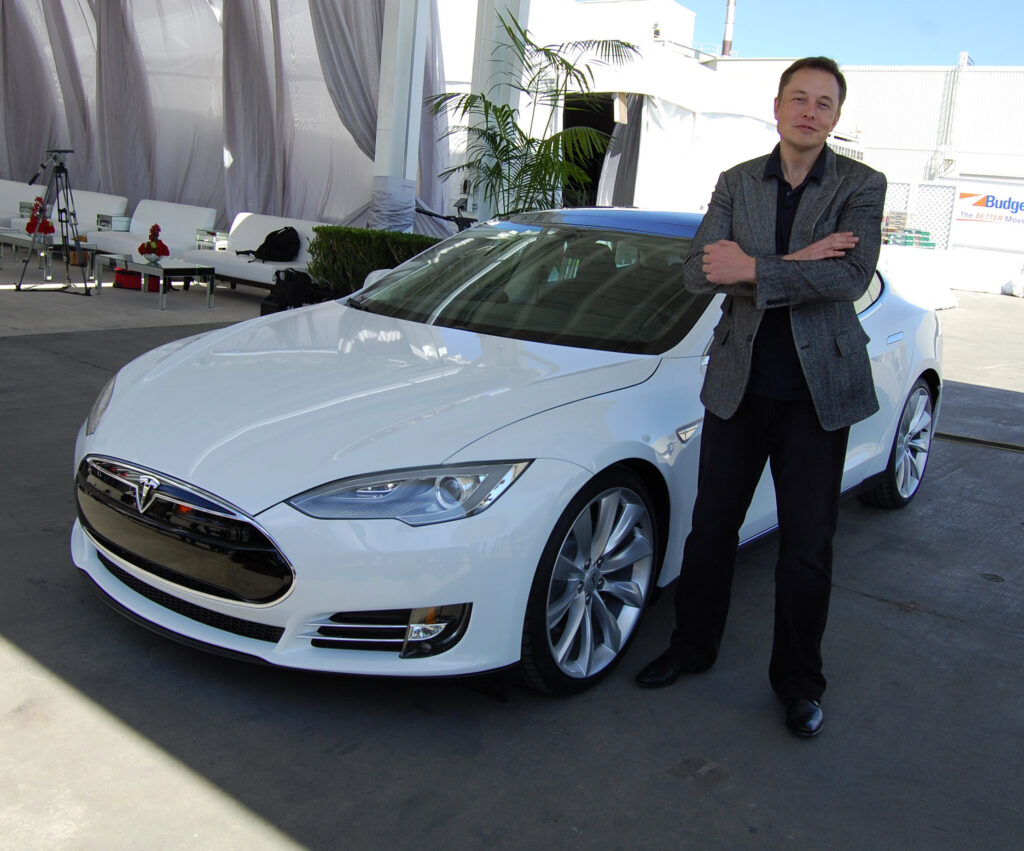 elonas muskas stovi salia tesla elektromobilio
