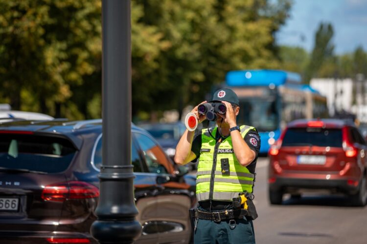 policijos pareigunas stebi eisma per ziuronus salia gatves vaziuojamosios kelio dalies
