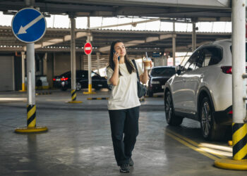 Automobilių stovėjimo aikštelėje telefonu kalbanti jauna moteris su kava