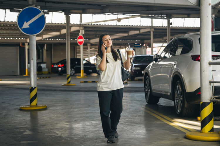 Automobilių stovėjimo aikštelėje telefonu kalbanti jauna moteris su kava