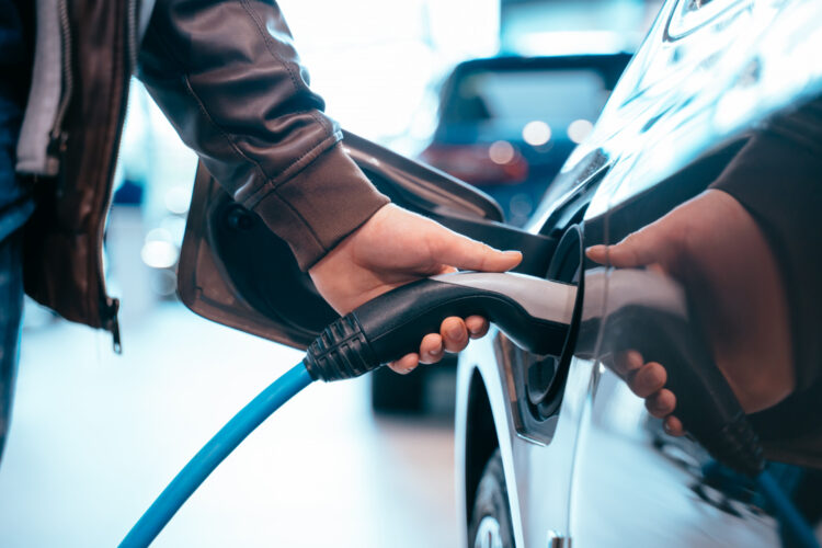 Žmogaus ranka laiko elektrinio automobilio įkrovimo jungtį prie elektrinio automobilio