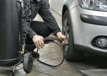 Vyras juodoje striukėje ir juodose kelnėse sėdintis šalia sidabrinės mašinos tikrina padangu slegi
