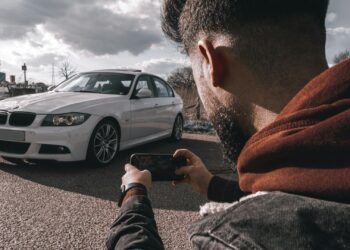 Vyras juodoje striukėje laikantis mobilųjį telefoną daro automobilio nuotrauka