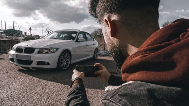 Vyras juodoje striukėje laikantis mobilųjį telefoną daro automobilio nuotrauka