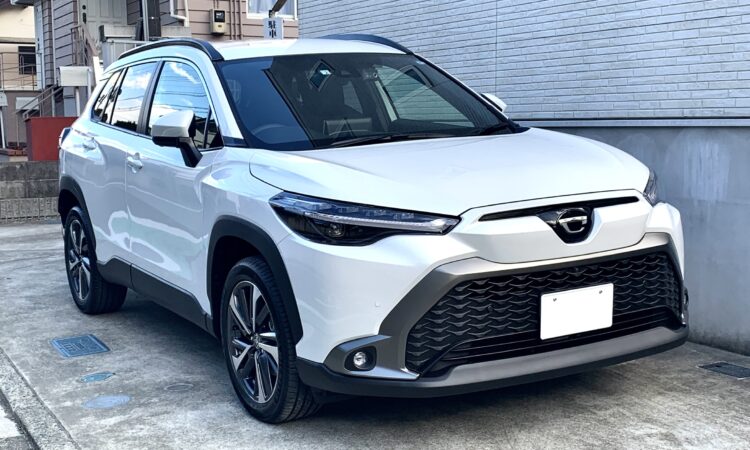 2022 Baltas Toyota Corolla Cross hibridinis visureigis iš priekio ir šono stovintis prie namo