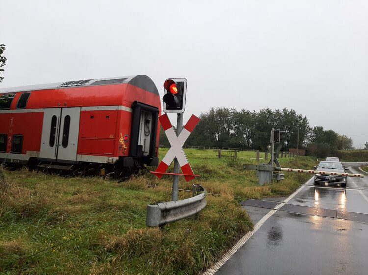 Degantis raudonas signalas prie geležinkelio pervažos, pro šalį važiuoja traukinys.