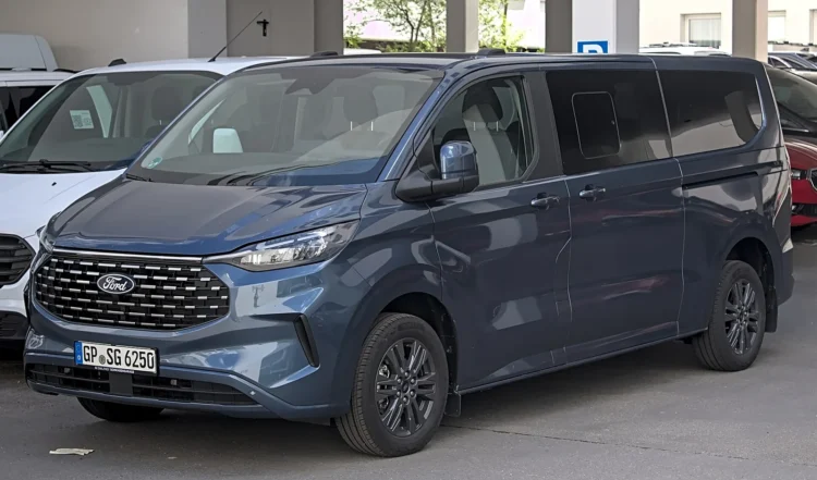 Daugiavaikėms šeimoms – septynvietis hibridas, su kuriuo sunkiam nameliui ant ratų neužteks B kategorijos 1 Ford Tourneo Custom: tamsiai mėlynas naujos kartos vienatūris aikštelėje, matomas priekis ir šonas